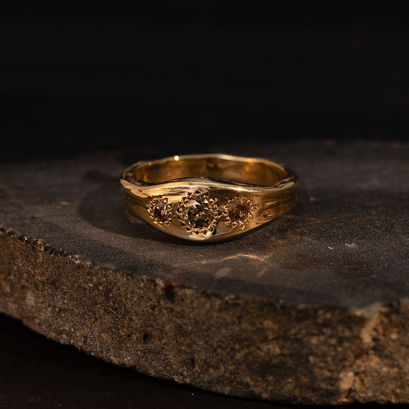 Molten Ring, Brandy Diamond, 9kt Yellow Gold - 9kt solid yellow gold ring with a subtle textured finish, featuring three round faceted brown diamond gemstones in a sunflower setting - displayed on black stone
