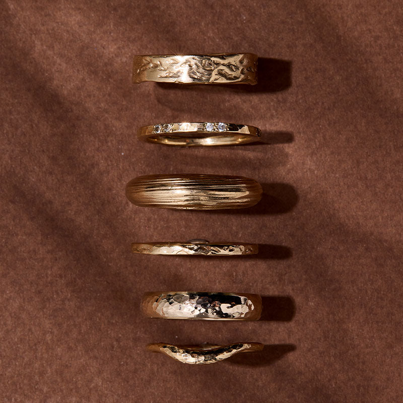 Textured Bands Stack – Six 9kt solid yellow gold bands in varying textures and widths, stacked atop brown paper. A branch-like shadow casts over them, accentuating their handcrafted details and warm, buttery golden hues.