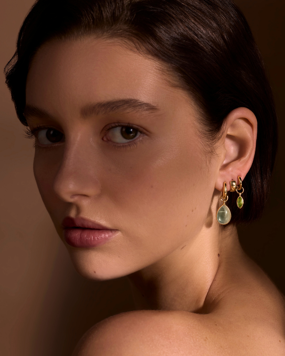 Close-up of a woman wearing gold earrings with green gemstones against a brown background