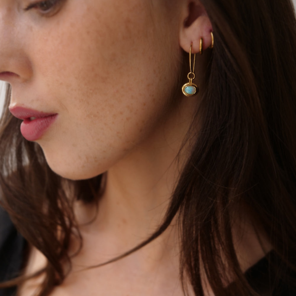 A close-up of a woman's ear wearing a delicate gold hook earring with a small oval light blue opal gemtone pendant.