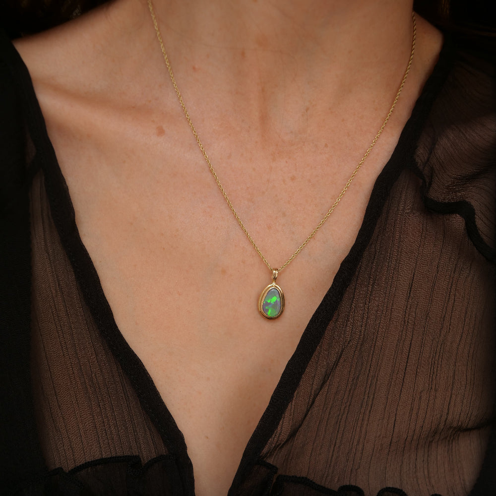 A close-up of a model wearing a delicate gold chain necklace with a teardrop-shaped green opal pendant, styled with a sheer black top.