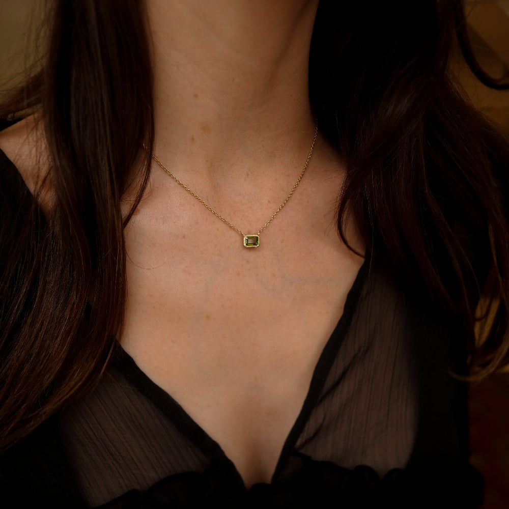 A close-up lifestyle shot of a woman wearing a delicate gold chain necklace with a small square green tourmaline gemstone pendant, styled with a black sheer top.