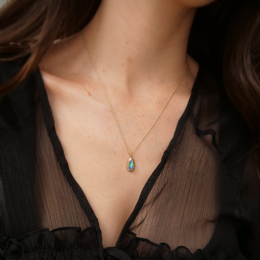 A close-up of a model wearing a delicate gold chain necklace with a teardrop-shaped iridescent opal pendant, styled with a black sheer top.