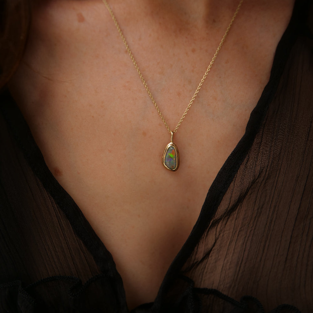 A gold chain necklace with a organic shaped iridescent opal pendant, worn by a model in a black sheer top.
