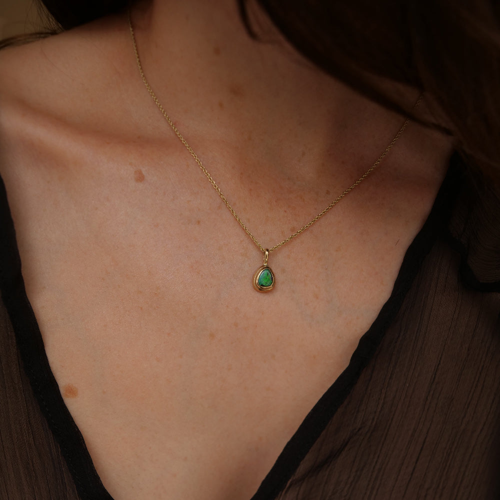 A close-up of a model's chest wearing a delicate gold chain necklace with a small teardrop-shaped opal pendant.