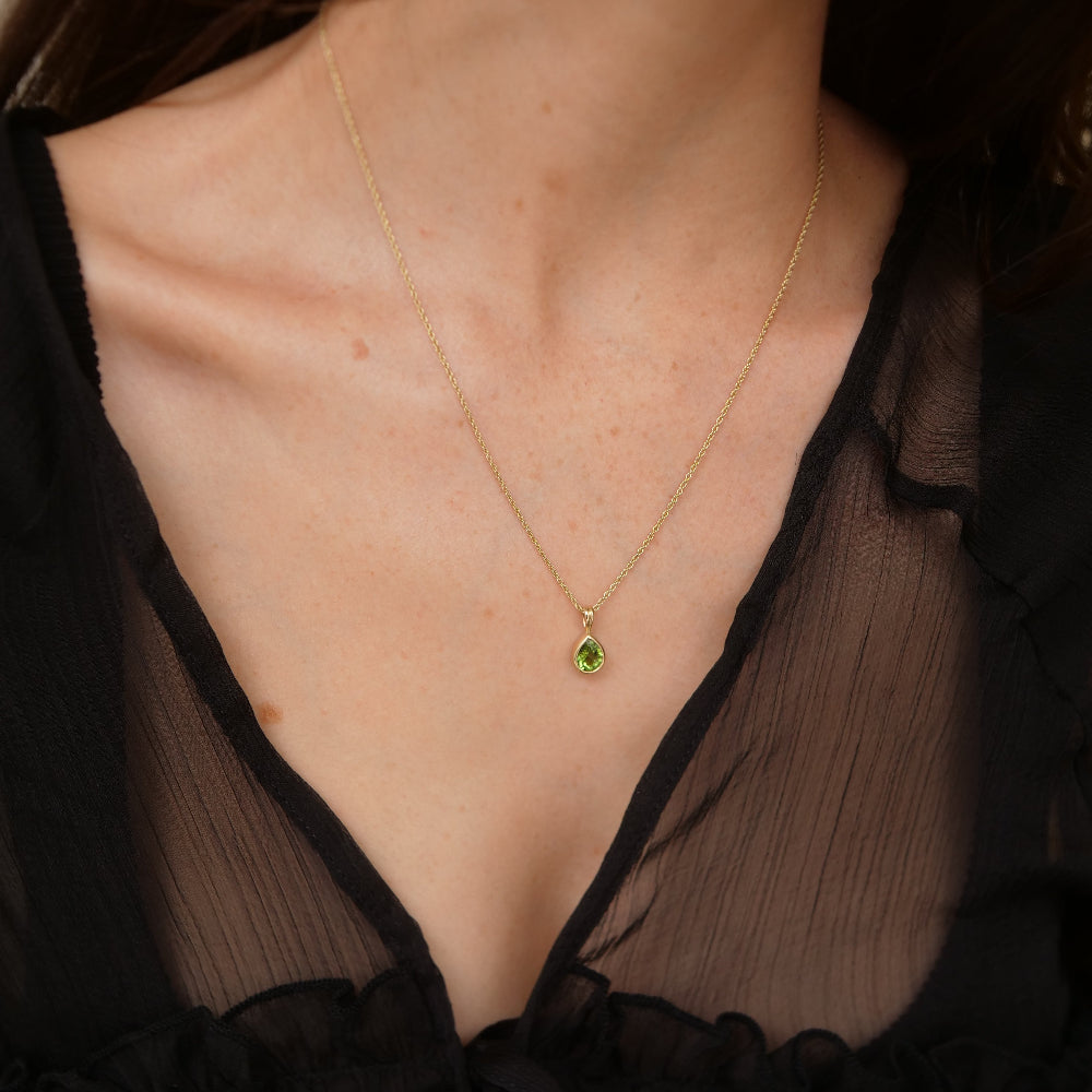 A close-up of a model wearing a delicate gold chain necklace with a small teardrop-shaped green tourmaline gemstone pendant, styled with a black sheer V-neck top.